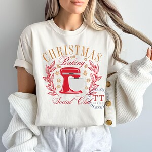 May include: White t-shirt with a red and gold design featuring a kitchen mixer and the text "Christmas Baking Social Club".