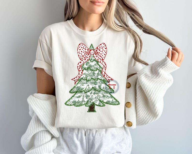 May include: A white t-shirt with a green Christmas tree design and a red and white polka dot bow. The tree is decorated with small red and white polka dots.