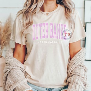 K&ouml;nnte beinhalten: Eine Frau tr&auml;gt ein hellbeiges T-Shirt mit dem Text "Outer Banks North Carolina" in rosa Schrift. Sie tr&auml;gt einen hellbraunen Cardigan und hat lange blonde Haare.