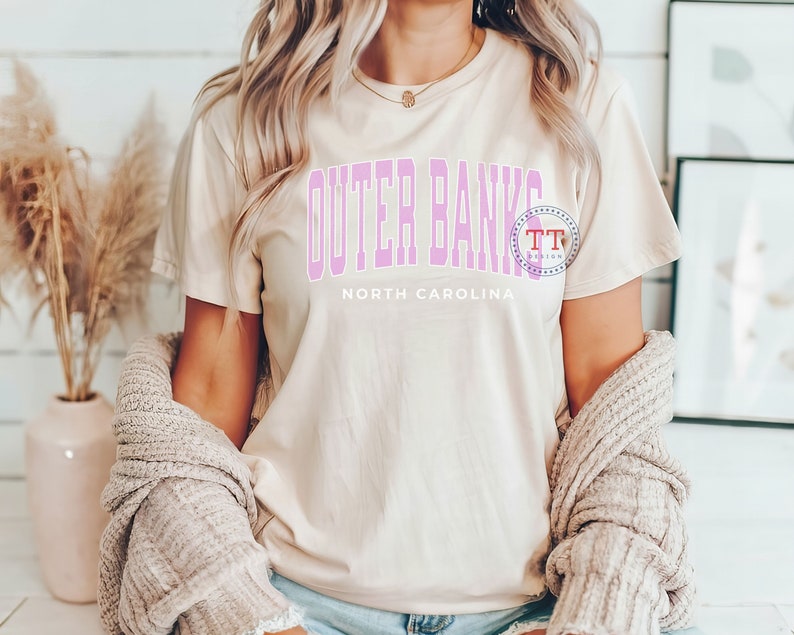K&ouml;nnte beinhalten: Eine Frau tr&auml;gt ein hellbeiges T-Shirt mit dem Text "Outer Banks North Carolina" in rosa Schrift. Sie tr&auml;gt einen hellbraunen Cardigan und hat lange blonde Haare.