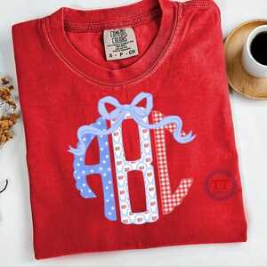 May include: Red Comfort Colors t-shirt with a patriotic design. The design features the letters "ABC" in a circle, with a blue bow and ribbon. The letters are decorated with stars, flags, and a red and white gingham pattern.