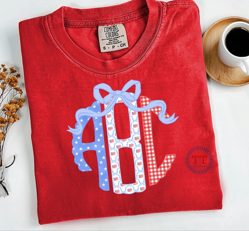 May include: Red Comfort Colors t-shirt with a patriotic design. The design features the letters "ABC" in a circle, with a blue bow and ribbon. The letters are decorated with stars, flags, and a red and white gingham pattern.