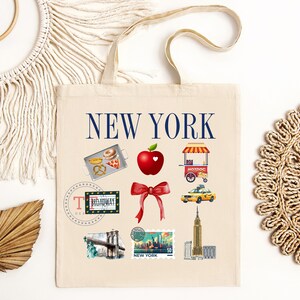Op de afbeelding: Een witte canvas tote bag met de tekst "NEW YORK" in blauwe letters. De tas heeft verschillende illustraties van bezienswaardigheden en iconen van New York City, waaronder een hotdogkraam, een gele taxi, de Brooklyn Bridge en een rode appel met een hart.