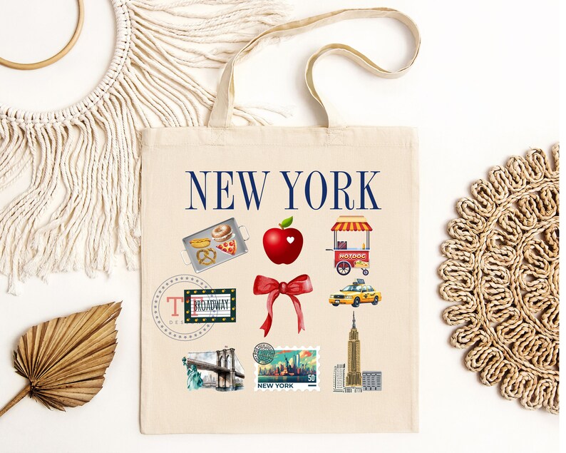 Op de afbeelding: Een witte canvas tote bag met de tekst "NEW YORK" in blauwe letters. De tas heeft verschillende illustraties van bezienswaardigheden en iconen van New York City, waaronder een hotdogkraam, een gele taxi, de Brooklyn Bridge en een rode appel met een hart.