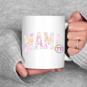 Puede incluir: Taza de cer&aacute;mica blanca con un dise&ntilde;o rosa y blanco con la palabra "MAMA" en una gama de colores pastel. El dise&ntilde;o incluye peque&ntilde;os conejos y flores de colores pastel.