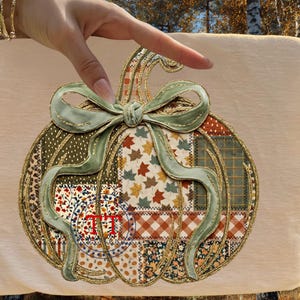 May include: A decorative pumpkin design with a patchwork pattern in various fabrics, including floral, polka dot, and plaid. The pumpkin features a green ribbon and gold trim. The design is on a beige background.