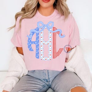 May include: Pink t-shirt featuring a monogram design with the letters ABC. The letter A is blue with white stars, B is white with red and blue patterns, and C is red gingham. A blue bow tops the design.