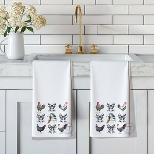 May include: Two white kitchen towels with a black and white checkered bow and chicken design. The towels are hanging on a white kitchen counter.