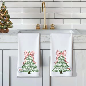 May include: Two white kitchen towels with a green Christmas tree design and a red and white polka dot bow on each tree. The towels are hanging on a white tiled backsplash.