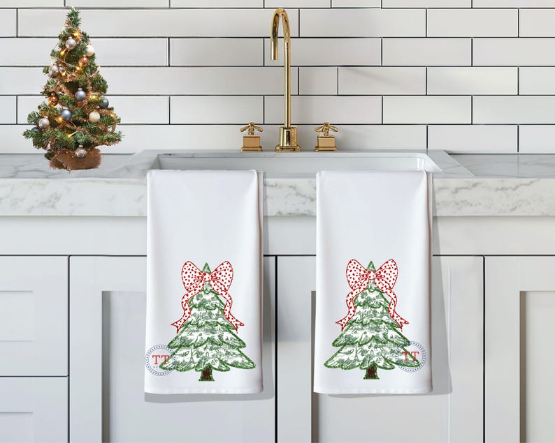 May include: Two white kitchen towels with a green Christmas tree design and a red and white polka dot bow on each tree. The towels are hanging on a white tiled backsplash.