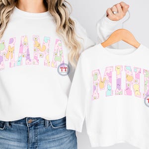Puede incluir: Dos camisas blancas de manga larga con las palabras "MAMA" y "MINI" escritas en letras rosas. Las letras est&aacute;n rellenas de caramelos Peeps de colores pastel. Las camisas son un conjunto a juego.