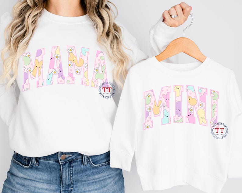 Puede incluir: Dos camisas blancas de manga larga con las palabras "MAMA" y "MINI" escritas en letras rosas. Las letras est&aacute;n rellenas de caramelos Peeps de colores pastel. Las camisas son un conjunto a juego.