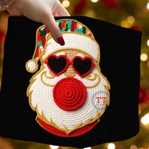 Op de afbeelding: Een zwart stoffen item met een gehaakt gezicht van de kerstman. De kerstman heeft hartvormige zonnebrillen, een rode neus en een witte baard en hoed met rode, groene en gouden accenten. Het ontwerp heeft een gouden omtrek en een cirkelvormig logo.
