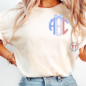 May include: Cream-colored t-shirt featuring a monogram design with the letters "ABC". The letters are decorated with blue polka dots, red gingham, and a blue bow. The shirt has short sleeves and a relaxed fit. The logo "TT DESIGN" is on the lower right.