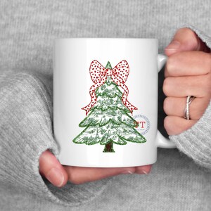 May include: White ceramic mug with a green Christmas tree design and a red and white polka dot bow. The tree is decorated with small, white dots.