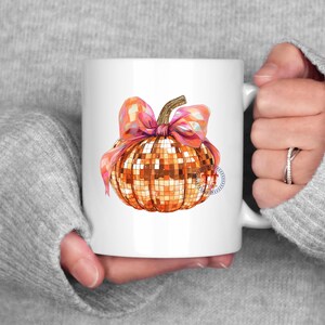 K&ouml;nnte beinhalten: Wei&szlig;e Keramiktasse mit einem goldenen Glitzerk&uuml;rbis mit einer rosa Schleife. Der K&uuml;rbis ist im Discokugel-Design.