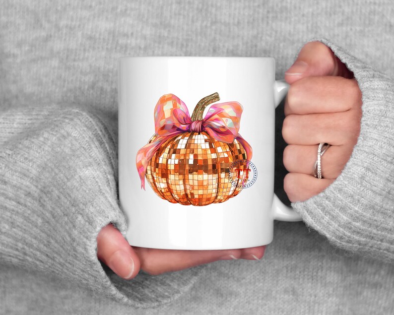 K&ouml;nnte beinhalten: Wei&szlig;e Keramiktasse mit einem goldenen Glitzerk&uuml;rbis mit einer rosa Schleife. Der K&uuml;rbis ist im Discokugel-Design.