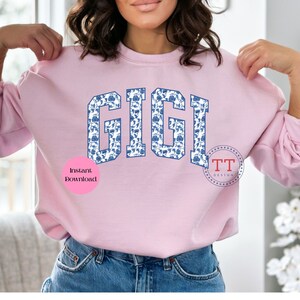 May include: A pink sweatshirt with the word "GIGI" printed in blue letters with a white and blue floral pattern. The letters are in a collegiate style font. The words "Instant Download" are printed in pink on a white circle below the word "GIGI".