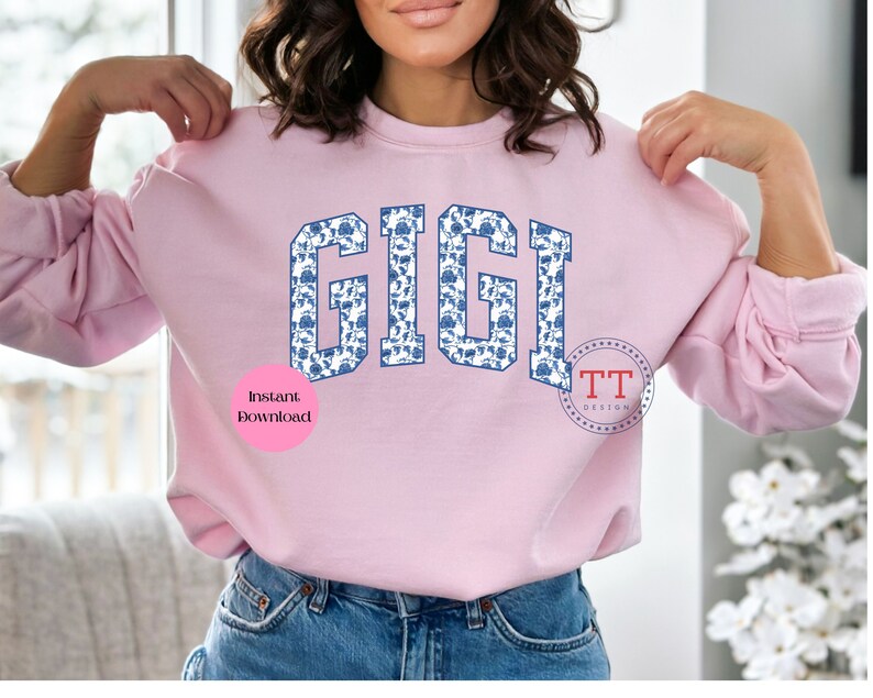 May include: A pink sweatshirt with the word "GIGI" printed in blue letters with a white and blue floral pattern. The letters are in a collegiate style font. The words "Instant Download" are printed in pink on a white circle below the word "GIGI".