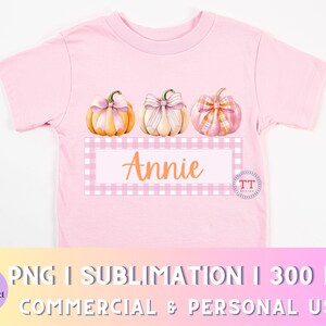 Puede incluir: Una camiseta rosa con un diseño de tres calabazas con lazos rosas y el nombre "Annie" en una caja a cuadros rosa y blanca.