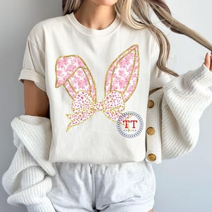 May include: A white t-shirt with a pink and gold bunny ear design. The ears are decorated with a floral pattern and a gold bow.