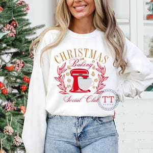 May include: White long-sleeved sweatshirt with a red and gold Christmas baking social club design. The design features a red stand mixer, a cup of coffee, and a wreath with gold stars and leaves.