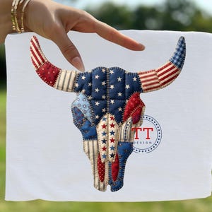 Puede incluir: Una tela cuadrada blanca con un diseño de calavera de toro. La calavera está compuesta de retazos en telas rojas, blancas y azules con estrellas y rayas. El diseño incluye el texto "TT DESIGN" en un emblema circular.