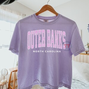 K&ouml;nnte beinhalten: Ein lavendelfarbenes T-Shirt mit dem Text "Outer Banks North Carolina" in rosa Schrift.