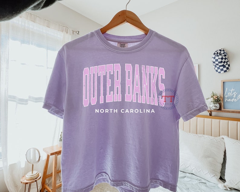 K&ouml;nnte beinhalten: Ein lavendelfarbenes T-Shirt mit dem Text "Outer Banks North Carolina" in rosa Schrift.