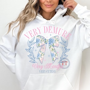 Peut inclure: Sweat-shirt blanc avec un n&oelig;ud rose et un motif floral. Le texte "Very Demure Very Mindful Very Cutesy" est imprim&eacute; sur le devant du sweat-shirt.