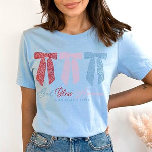 Puede incluir: Una camiseta azul claro con tres lazos rojos, rosas y azules con un patr&oacute;n floral. La camiseta tiene el texto "God Bless America" y "Land That I Love" impreso en ella.