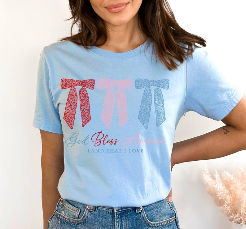 Puede incluir: Una camiseta azul claro con tres lazos rojos, rosas y azules con un patr&oacute;n floral. La camiseta tiene el texto "God Bless America" y "Land That I Love" impreso en ella.