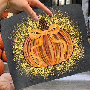 May include: A dark gray t-shirt featuring a hand-painted pumpkin design. The pumpkin is orange and yellow with a bow. The background has yellow speckles. Pumpkins are in the background, suggesting a fall or Halloween theme.
