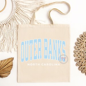 May include: A beige canvas tote bag with blue text that reads "Outer Banks North Carolina". The text is in a stylized font and has a blue circle with the letters "TT" in it.