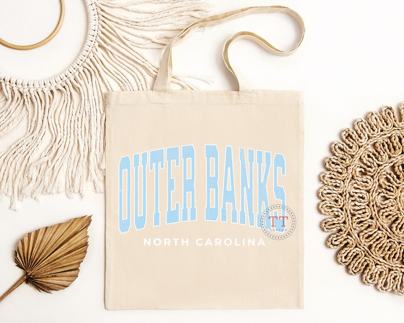 May include: A beige canvas tote bag with blue text that reads "Outer Banks North Carolina". The text is in a stylized font and has a blue circle with the letters "TT" in it.