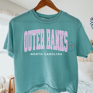 K&ouml;nnte beinhalten: Ein blaues, &auml;rmelloses T-Shirt mit dem Text "Outer Banks" in rosa Schrift und "North Carolina" in wei&szlig;er Schrift.