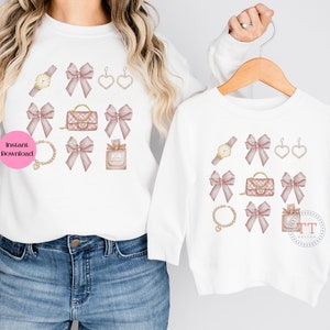 May include: Two white t-shirts with a pink bow and gold accessories design. The design includes a watch, earrings, a purse, a bracelet, and perfume bottle.
