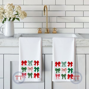 May include: Two white kitchen towels with red and green bows printed on them. The towels are hanging on a white kitchen cabinet.