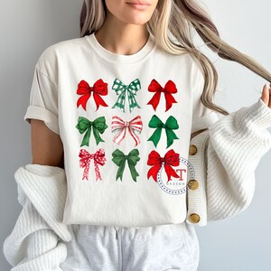 May include: White t-shirt with a design of nine red and green bows. The bows are arranged in three rows of three. The shirt has a crew neck and short sleeves.