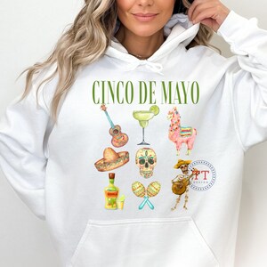 Pu&ograve; includere: Felpa con cappuccio bianca con una grafica verde "Cinco de Mayo". La grafica include una chitarra, una margarita, un lama, uno scheletro di zucchero, una bottiglia di tequila, maracas e uno scheletro che suona la chitarra.