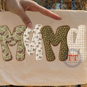May include: A tan-colored sign with the word "MAMA" in large, textured letters. The letters have different patterns: camouflage, ducks, and a woven texture. A blue and red circular logo with the text "TT DESIGN" is in the bottom right corner.