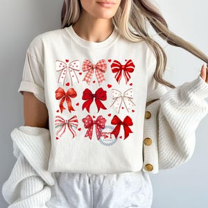 May include: White t-shirt with a red and white bow design. The bows are arranged in a grid pattern and have a heart design in the center.
