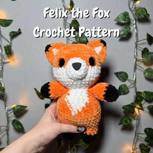 May include: A handmade orange, white, and black crochet fox plushie. The fox has a white chest, black paws and ears, and large black eyes. Text on the image reads "Felix the Fox Crochet Pattern".