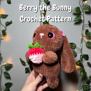 May include: A brown crochet bunny holding a crochet strawberry. The bunny has large black eyes with pink accents and a pink bow. The text "Berry the Bunny Crochet Pattern" is at the top of the image.