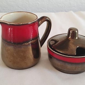 May include: A ceramic creamer pitcher and a matching lidded sugar bowl. Both pieces feature a glossy finish with a vibrant red band around the upper section, transitioning into a metallic bronze and brown glaze on the lower body and handle.