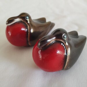May include: Two ceramic bird figurines shaped like robins, each featuring a glossy, bright red chest and a dark brown, smooth body. The birds have small, dark eyes and a rounded, compact form, positioned side by side on a light-colored surface.