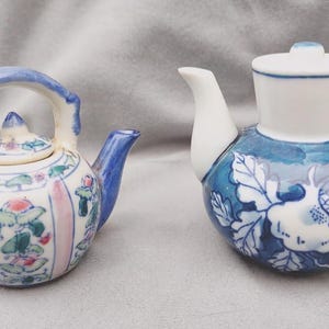 May include: Two small ceramic teapots. The left teapot features a white base with vertical pink stripes, blue handle and spout, and floral patterns. The right teapot has a round body with blue and white floral designs and a solid white spout and lid.