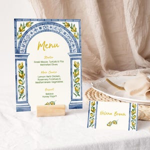 May include: A rectangular menu card and a smaller place card featuring a blue and white tile-patterned archway with yellow lemon illustrations. The menu lists courses including Greek Mezze, Lemon Herb Chicken, and Baklava. The name Helena Brown is printed on the place card. Both cards are displayed on a wooden block.