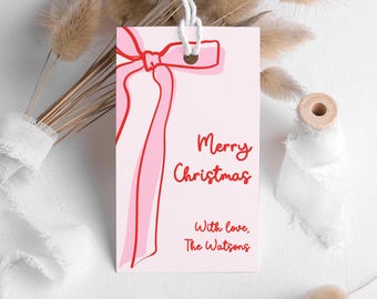 Può includere: Un'etichetta regalo rosa chiaro con un'illustrazione di fiocco rosso e le parole "Merry Christmas" in corsivo rosso. Sotto c'è scritto "With love, The Watsons". L'etichetta è legata con uno spago bianco.