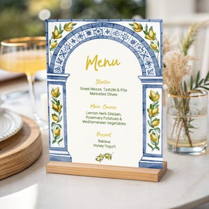 May include: A rectangular menu card standing in a light wood base, featuring a watercolor illustration of a blue tiled archway decorated with yellow lemons and green leaves. The text reads Menu, Starter, Greek Mezze, Tzatziki & Pita, Marinated Olives, Main Course, Lemon Herb Chicken, Rosemary Potatoes & Mediterranean Vegetables, Dessert, Baklava, Honey Yogurt.